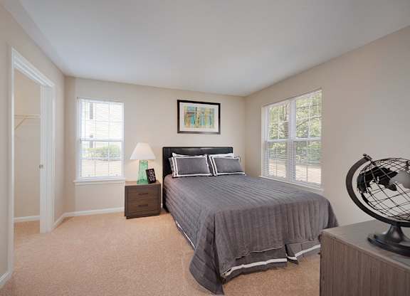 An apartment bedroom with carpeting shows a bed with windows on both sides, a night stand, and a glimpse of a dresser.