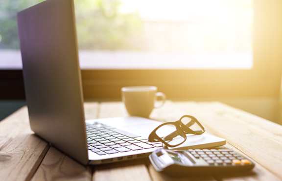 A laptop, a pair of glasses, a calculator, and a cup are placed on a wooden table.