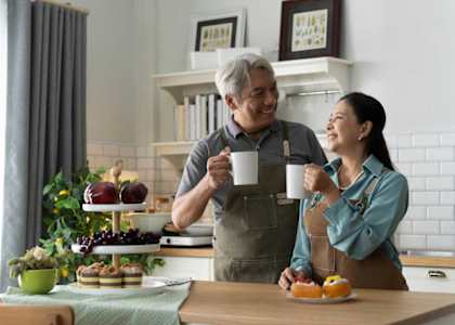 A man and woman are standing in a kitchen, both holding coffee mugs and smiling at each other.