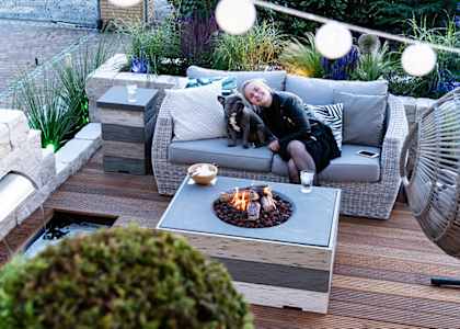 A woman is sitting on a grey couch with a dog beside her, in front of a fire pit.