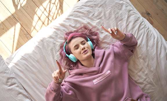 A person with pink hair is lying on a bed and wearing headphones.