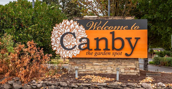 A sign that says "Welcome to Canby the garden spot" is displayed in front of a stone wall.
