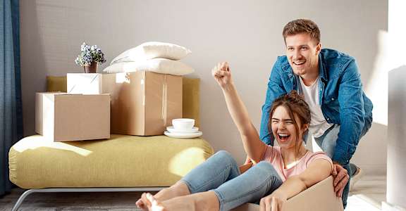 A man is lifting a woman out of a cardboard box in a room with boxes and a couch.