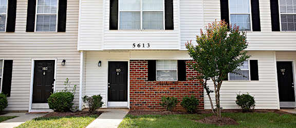 A white house with black shutters and a brick wall in front.