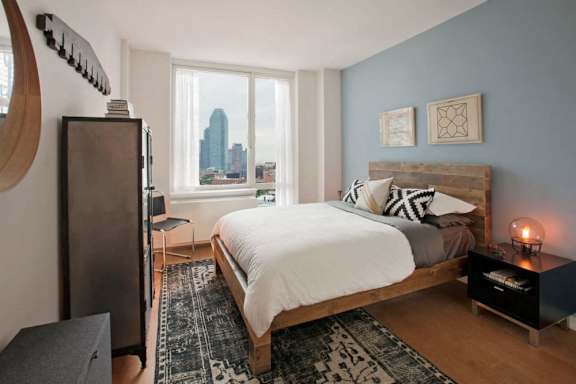 A bedroom with a large bed and a city view through the window at The Maximilian Apartments, Long Island City