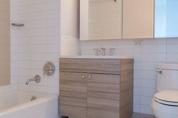 A white toilet sits next to a sink with a wooden cabinet above it at The Maximilian Apartments, Long Island City, 11101