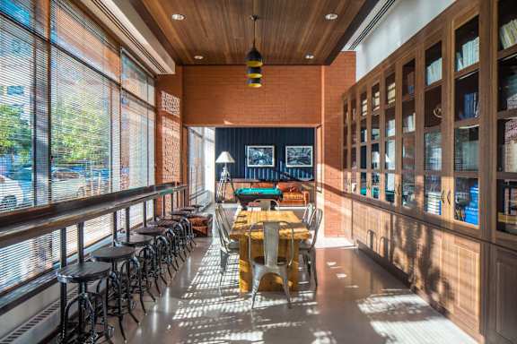 A long wooden table is surrounded by chairs in a room with a brick wall and a long window at The Maximilian Apartments, Long Island City, 11101