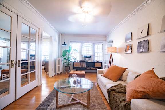 A living room with a glass table and a couch with orange pillows.