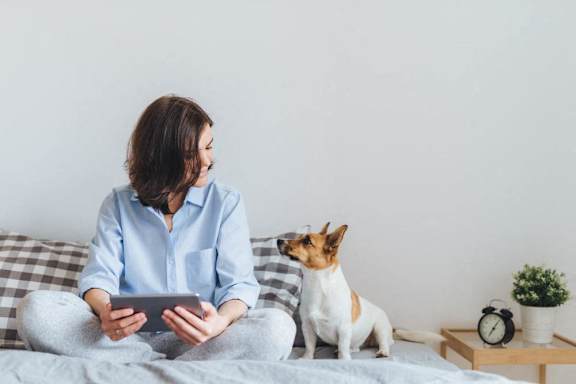 a woman sitting on her bed with her dog holding a tablet