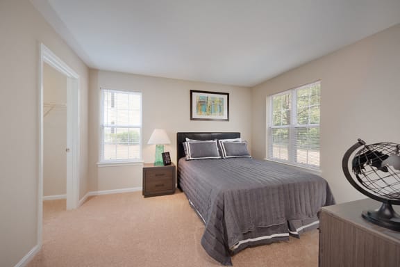An apartment bedroom with carpeting shows a bed with windows on both sides, a night stand, and a glimpse of a dresser.