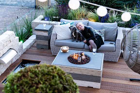 A woman is sitting on a grey couch with a dog beside her, in front of a fire pit.