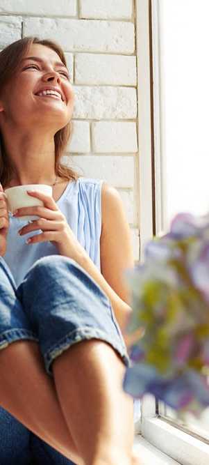 a woman sitting on a window sill laughing and holding a cup of coffee