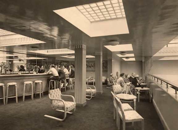 A black and white photo of a diner with people sitting at tables and a bar area.