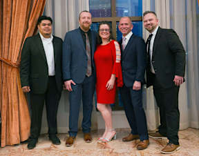 Five people in suits and dresses stand in front of a window.