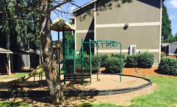 A playground with a green swing set and a tree in the foreground. at Fort Vancouver Terrace, Vancouver, WA, 98661