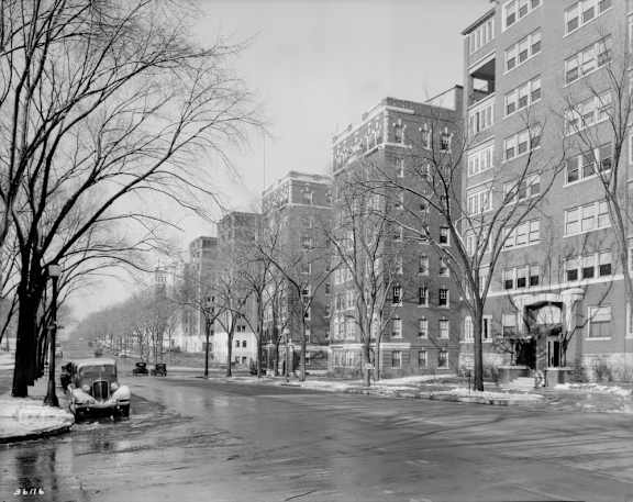 A black and white photo of a snowy street with cars and buildings.