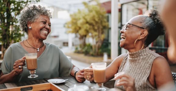 Two women laughing and drinking coffee outside.
