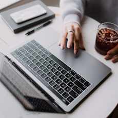 A person is typing on a laptop with a glass of drink on the side.