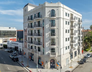 A large white building with balconies is situated on a street with cars parked on the side.