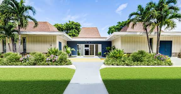 Entrance to the leasing office surrounded by palm trees and landscaped lawns at Coronado Springs Apartments in Palm Springs, FL. Pet-friendly community with one- and two-bedroom apartments.