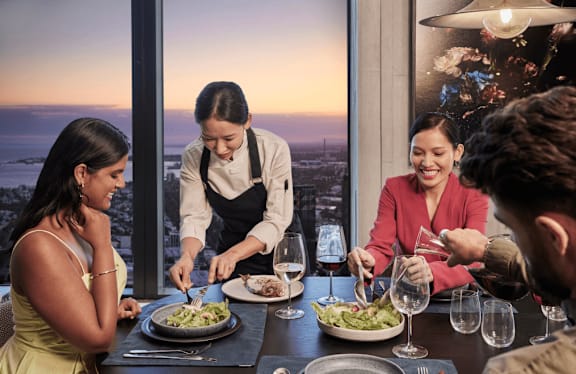 A waiter is serving food to a woman at a restaurant with a view of the city.