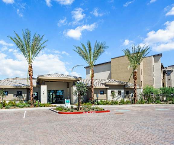 The exterior of the tan front office building and the parking lot with palm trees and plants at Reflect at Dobson Ranch Apartment Homes. 