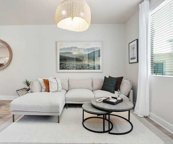 The cozy, white living room with a white couch, coffee table, a mirror, overhead lighting, decorations, a TV, and window at Reflect at Dobson Ranch Apartment Homes. 