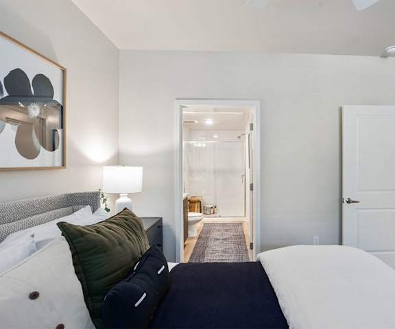 A large/cozy bedroom with a queen-size bed, a dresser, decorations, window, and nightstands with a view of the bathroom and a door to the kitchen at Reflect at Dobson Ranch Apartment Homes. 
