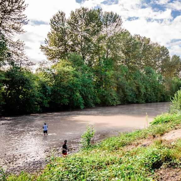 Two people are wading through a river in a natural landscape.