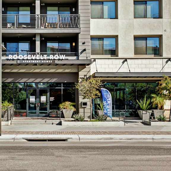 The front entrance to Roosevelt Row Apartments, showing the front sign, trees lining the street, and a cafe with dining to the left. 