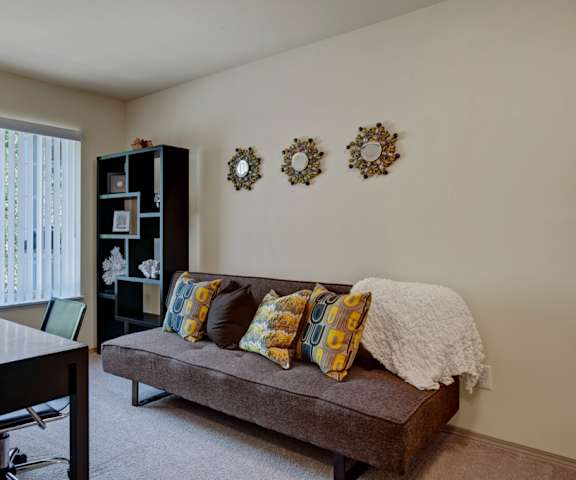 An office with a desk, chair, orchid, plush couch with throw blanket and pillows, wall art, shelves with artwork and other decorations, and windows with blinds at Windsong Apartment Homes. 