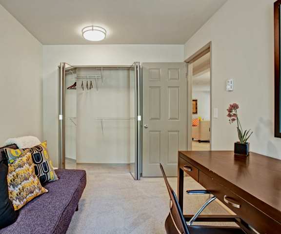 An office with a desk, chair, orchid, plush couch with throw blanket and pillows, wall art, shelves with artwork and other decorations, and a closet with mirrored doors and hangers at Windsong Apartment Homes. 