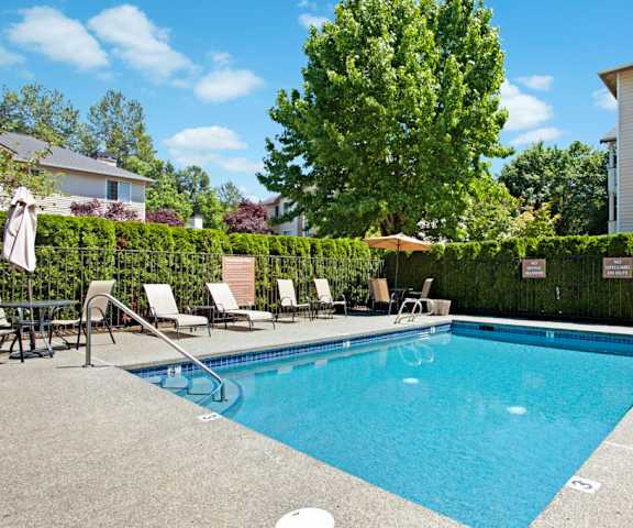 The large, turquoise outdoor pool with pool chairs, tables, and chairs with the apartments in the background at Windsong Apartment Homes. 