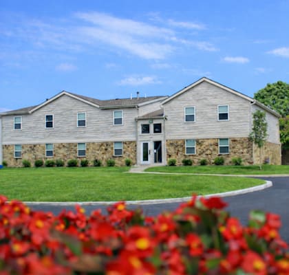 A building with a grey stone facade and a white door is surrounded by a green lawn and red flowers.