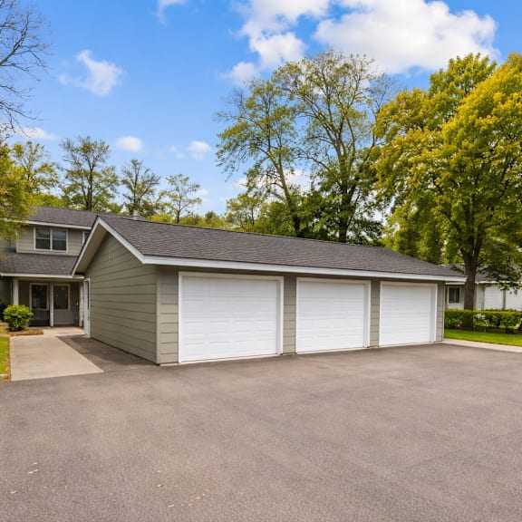A two-car garage with a grey roof and white doors is surrounded by a driveway and trees.