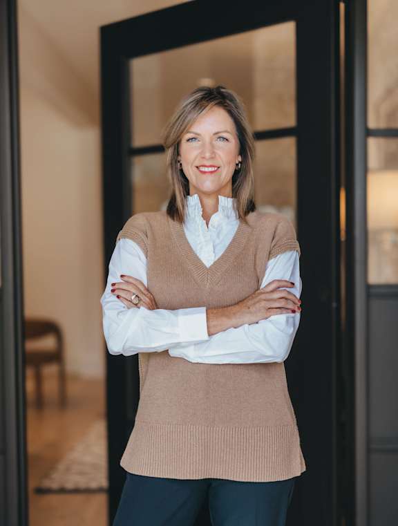 A woman with her arms crossed wearing a tan vest and white shirt.