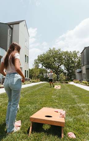 A woman in a white top and blue jeans stands on a lawn with a wooden table in front of her.