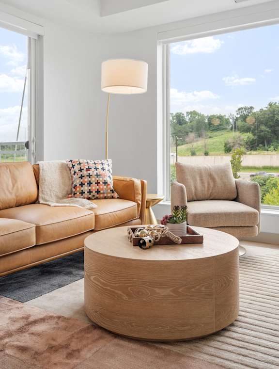 A living room with a tan couch, a round coffee table, and a large window.
