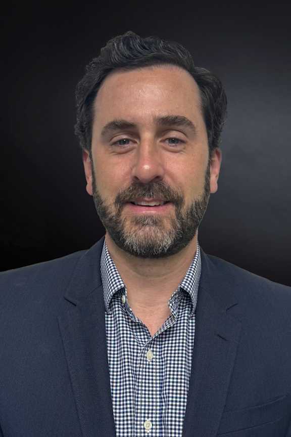 A man with a beard and wearing a checkered shirt under a suit jacket.