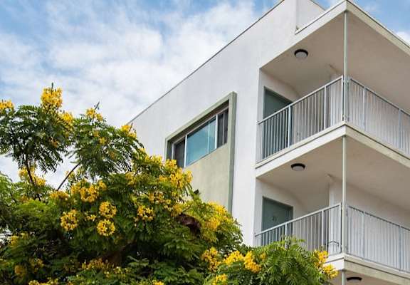 A white building with balconies and windows is surrounded by greenery.