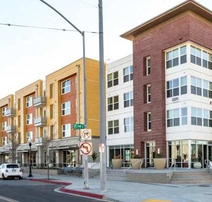 A multi-story building with a red brick facade is situated on a street corner with cars parked on the side.