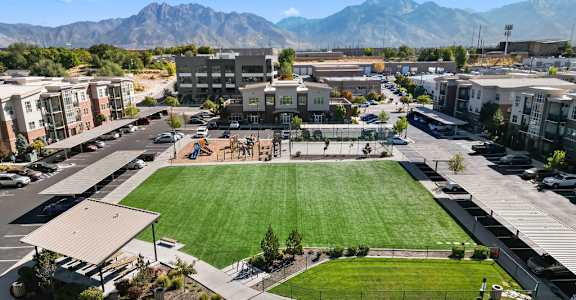 A large grassy area in the middle of a parking lot with buildings and mountains in the background.