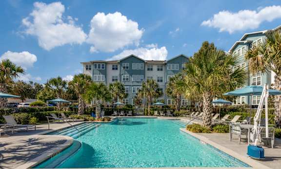 A large swimming pool with a blue tinted water surrounded by palm trees and a white building in the background.