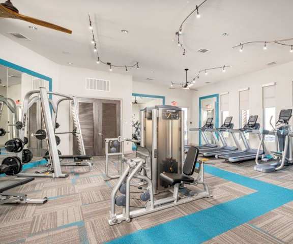 A spacious gym with cardio machines, ceiling fans, blue accents, windows, TVs, and weights at the Flats at San Tan Apartment Homes. 