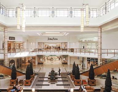 The image shows a spacious indoor shopping mall with a variety of stores and a central atrium with seating and decorative trees.