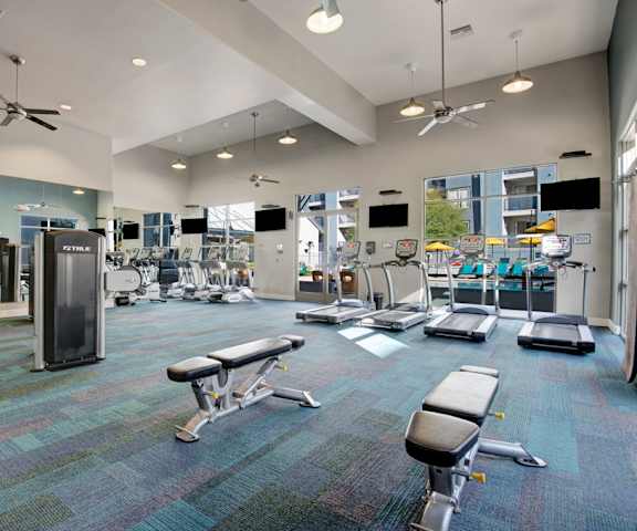 A spacious gym with cardio machines, windows, TVs, and weights at Rio Paradiso Apartment Homes. 