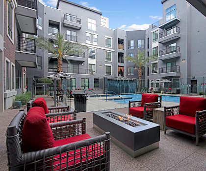 The outdoor lounge area with plush red chairs, a firepit, pool chairs and umbrellas, trees, and the pool with a waterfall at Park on Central Apartments. 