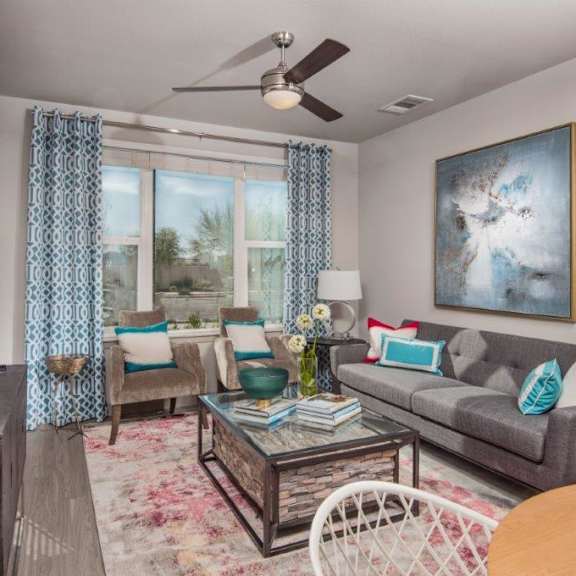A living room at Escape Apartment Homes with a gray couch, light brown plush velvet chairs, a glass coffee table, artwork, a TV, a large window with blue patterned curtains, a ceiling fan with a light, and lamps at the Flats at San Tan Apartment Homes. 