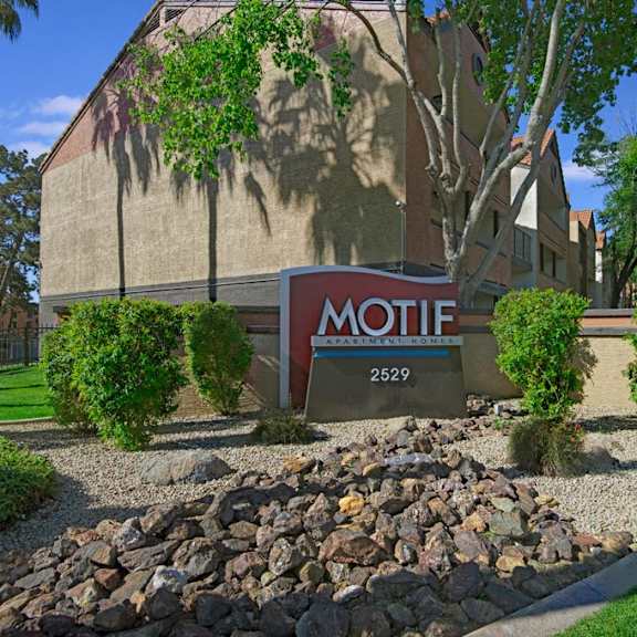 Entrance to the apartment complex Motif with a sign surrounded by shrubs and trees in front of an apartment building. 