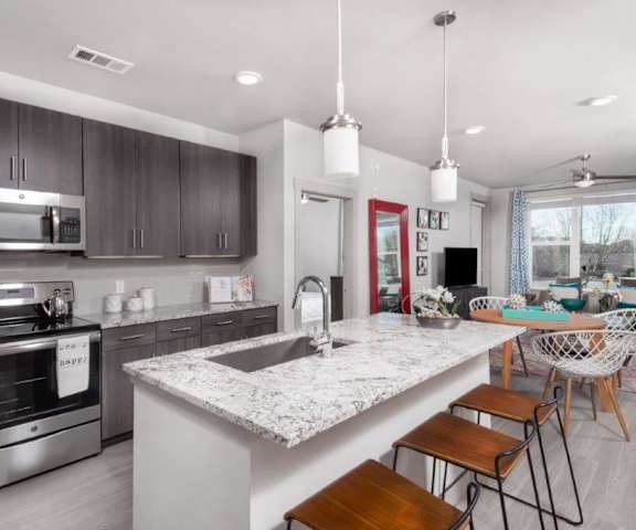 A large, modern kitchen with stainless steel appliances, kitchen utensils, marble countertops with lots of room, a kitchen island with three barstools, and large, dark wooden cabinets at the Flats at San Tan Apartment Homes. 